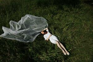 Mañana de el novia en naturaleza. el novia es en un vendaje vestido y un largo velo, acostado en el verde césped y disfrutando el del sol calor. amplio ángulo foto. foto