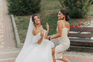 Wedding photo in nature. A brunette bride in a white long dress and her friend in a nude dress are fooling around together and smiling sincerely. Young women. emotions