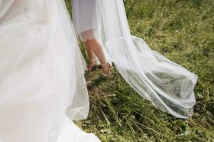 De las mujeres piernas en Boda zapatos, en el antecedentes de verde césped y un Boda vestido. detalles. Boda accesorios. un velo estropeado por el viento. primavera boda. foto