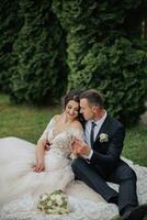 retrato de el novia en naturaleza. un morena novia en un blanco cordón vestir y un elegante novio en un negro traje son sentado en el césped, posando, participación un ramo. hermosa constituir. abierto espalda. foto