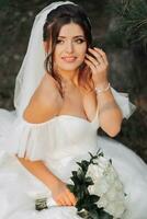 portrait of the bride in nature. A brunette bride in a white voluminous dress is sitting, posing near a coniferous tree, holding a bouquet of white roses. Beautiful hair and makeup. Warm light photo