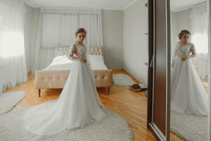 Preparation for the wedding. Beautiful young bride in white wedding dress indoors. A luxurious model looks at the mirror at home in a studio room with a large window. The girl shows photo