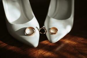 Photo of details at the wedding. Wedding rings of the bride and groom stand on white classic shoes, which are located on a wooden background. Contour line. Shadows and light.