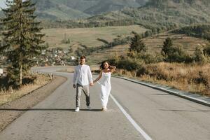 A stylish groom in a white shirt and a cute brunette bride in a white dress are walking on an asphalt road against the background of a forest and mountains. Wedding portrait of newlyweds. photo