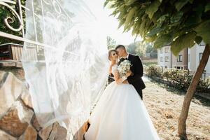 Wedding portrait. A groom in a black suit and a blonde bride stand embracing by a stone wall under a tree. A white, long veil in the air. Photo session in nature. Beautiful hair and makeup