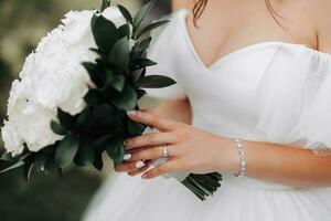 Cropped photo. The bride is wearing a white dress with a neckline, holding a bouquet of white roses. Beautiful hands and silver jewelry. French manicure. Portrait of the bride. photo