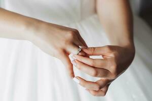 A brunette bride in a white dress puts on her silver wedding ring. French manicure. Open shoulders. Beautiful hands. Long veil. Morning of the bride. Details photo