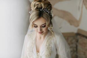 A bride with an elegant hairdo and tiara, in a robe, stands looking down and holding a snow-white veil against a background of painted walls. Beautiful makeup. Wedding photo