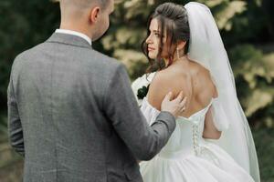 Portrait of the bride and groom in nature. A brunette bride in a long dress with open shoulders and a bouquet of roses stands with her shoulders to her groom, he touches her open shoulders. Stylish photo