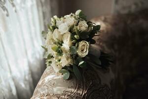 Accessories for the groom's wedding. A bouquet of white roses and greenery stands on the back of the sofa near the window. Beautiful flowers. Light and shadows photo