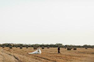 Wedding portrait of the bride and groom. Bride and groom on the background of hay, walk across the field to meet each other. Red-haired bride in a long dress. Stylish groom. Summer. Bales of hay photo