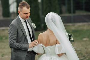 Portrait of the bride and groom in nature. A brunette bride in a long dress with open shoulders and a bouquet of roses stands with her shoulders to her groom, he touches her open shoulders. Stylish photo