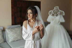 Brunette bride in a robe, standing in front of a mannequin with a dress, posing for the camera, looking to the side. Open shoulders. Elegant hairstyle. Voluminous veil. Morning of the bride photo
