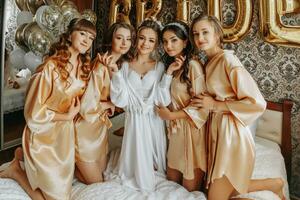 Young bridesmaids in silk robes in a hotel room. Beautiful women celebrating a bachelorette party sitting on the bed in themed pajamas. photo