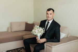 retrato. un hombre en un blanco camisa, negro Corbata y negro clásico traje se sienta en un marrón sofá con un ramo de flores de blanco rosas. un elegante mirar. de los hombres estilo. moda. negocio foto