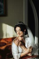 Portrait of a brunette bride sitting in a petticoat and touching her shoulder. The silver crown is decorated with steel flowers. Gorgeous make-up and hair. Voluminous veil. photo