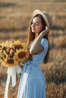 retrato de un joven muchacha. un niña en un azul vestir y sombrero, participación un ramo de flores de girasoles, en contra un antecedentes de heno fardos largo Derecho cabello. bonito color. verano foto