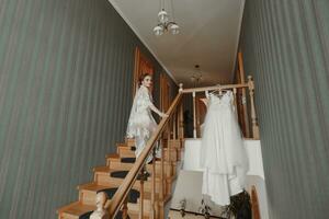 A delicate wedding dress hangs on a beautiful railing in the hallway of the house. The bride climbs the steps in a delicate transparent petticoat. Nice light. Wedding photo. A high quality photo