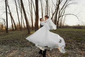 Wedding photo. The bride and groom are walking in the forest. The groom spins the bride in his arms, sincere smiles. Long wedding dress. A couple in love among tall trees. Autumn sunlight. photo