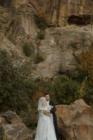 The bride and groom are standing on a rock high in the mountains next to a large stone. The bride is under a veil, the groom is hugging her shoulders photo