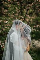 Happy young couple. Wedding portrait. The bride and groom gently snuggled up to each other under a veil against the background of a blooming bush. Wedding bouquet. Spring wedding photo