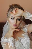 A blonde bride in a long, lacy petticoat is standing in her room, posing by the window, putting on her earrings. Beautiful hair and makeup, open bust. Wedding portrait in a light tone photo