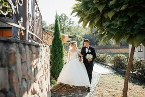 Wedding portrait. A groom in a black suit and a blonde bride walk holding hands near a stone wall under a tree. A white, long veil in the air. Photo session in nature. Beautiful hair and makeup