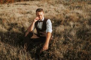 portrait of a stylish groom on a background of dry autumn grass. the concept of a rural wedding in the mountains, happy bohemian newlyweds. man relaxing sitting on the grass photo