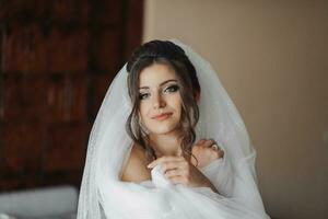 A brunette bride in a white dress, posing, wrapped in her voluminous veil. Open shoulders. Beautiful hands. Long veil. Morning of the bride. Portrait photo