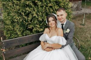 retrato de el novia y novio en naturaleza. un elegante novio y un morena novia en un blanco largo vestir sentar en un banco en contra un árbol fondo, abrazo y atentamente sonrisa. Rizado pelo foto