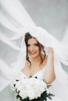 Curly brunette bride in a white dress, covered with a veil, poses for the camera with a bouquet of roses. Portrait of the bride. Beautiful makeup and hair. Wedding in nature photo