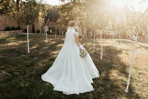 A blonde bride in a white dress with a train and lace sleeves holds a bouquet and stands with her shoulders to the camera. A long veil falls in the air. Photo session in nature. Sun rays