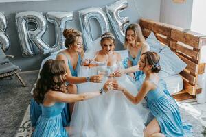 joven damas de honor en azul seda vestidos bebida champán en el de la novia habitación. hermosa mujer celebrando despedida de soltera fiesta sentado en cama y con champán. foto