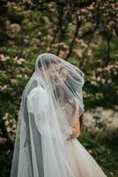 Happy young couple. Wedding portrait. The bride and groom gently snuggled up to each other under a veil against the background of a blooming bush. Wedding bouquet. Spring wedding photo
