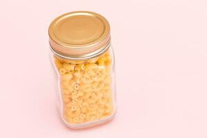 Uncooked Chifferi Rigati Pasta in Glass Jar on Pink Background. Fat and Unhealthy Food. Scattered Classic Dry Macaroni. Italian Culture and Cuisine. Raw Pasta photo