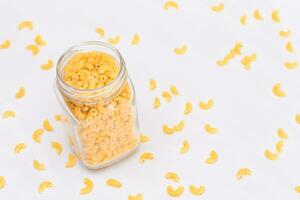 Uncooked Chifferi Rigati Pasta in Glass Jar on White Background. Fat and Unhealthy Food. Scattered Classic Dry Macaroni. Italian Culture and Cuisine. Raw Pasta photo