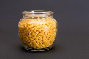 Uncooked Chifferi Rigati Pasta in Glass Jar on Black Background. Fat and Unhealthy Food. Classic Dry Macaroni. Italian Culture and Cuisine. Raw Pasta photo