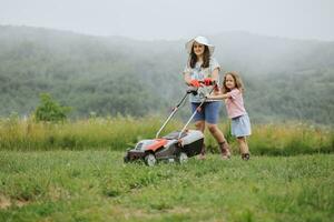 A woman in boots with her child in the form of a game mows the grass with a lawnmower in the garden against the background of mountains and fog, garden tools concept, work, nature. photo