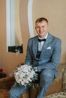 novio con un ramo de flores de flores en un habitación con un maravilloso interior. el novio mira dentro el lente foto