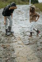 dos contento pequeño muchachas de europeo apariencia jugando en charcos durante lluvia en verano. niños son jugando en el lluvia. niño jugando en naturaleza al aire libre. el niña disfruta el lluvia. foto