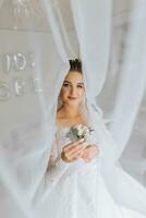 A brunette bride in a white dress holds her wedding bouquet, poses. French manicure. Open shoulders. Beautiful hands. Long veil. Morning of the bride. Details photo