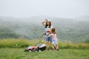 A woman in boots with her child in the form of a game mows the grass with a lawnmower in the garden against the background of mountains and fog, garden tools concept, work, nature. photo