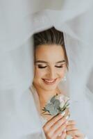 A brunette bride in a white dress holds her wedding bouquet, poses. French manicure. Open shoulders. Beautiful hands. Long veil. Morning of the bride. Details photo