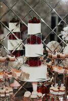 The white wedding cake is decorated with red roses. A delicious wedding. Candy bar for a banquet. Celebration concept. Fashionable desserts. A table with various sweets, candies. Fruits, cakes photo