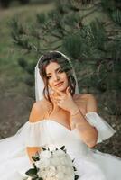 portrait of the bride in nature. A brunette bride in a white voluminous dress is sitting, posing near a coniferous tree, holding a bouquet of white roses. Beautiful hair and makeup. Warm light photo