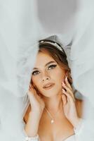 Beautiful curly brown-haired bride in a white dress poses for a photographer, standing under a veil in a beautiful dress with sleeves. Wedding photography, close-up portrait, chic hairstyle. photo