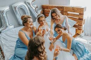 joven damas de honor en azul seda vestidos bebida champán en el de la novia habitación. hermosa mujer celebrando despedida de soltera fiesta sentado en cama y con champán. foto