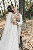 Wedding photo in nature. The bride hugs and kisses the groom. The bride has her shoulders turned. Beautiful hairstyle and hair decoration. Portrait of the bride and groom in the forest