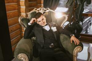 portrait of positive young groom in black suit with white shirt smiling and sitting on comfortable dark green sofa in modern hotel room on wedding day photo