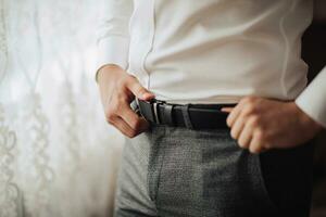 detalles. un hombre en un blanco camisa y gris pantalones ajusta su elegante cinturón. masculino estilo. moda. negocio foto